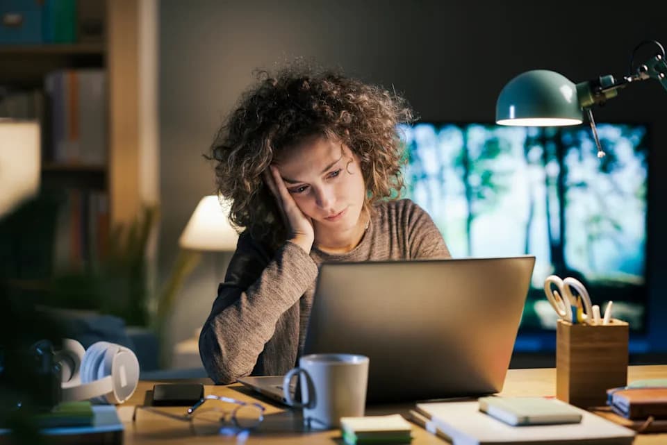 A woman looks intently at a laptop while working from a home office at night, illustrating online work-from-home scams.